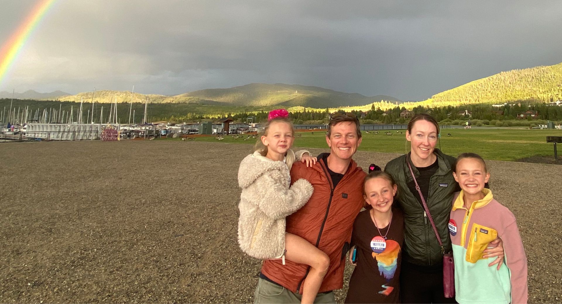 Family of five smiling on a lakeshore beneath a bright double rainbow and mountain sky