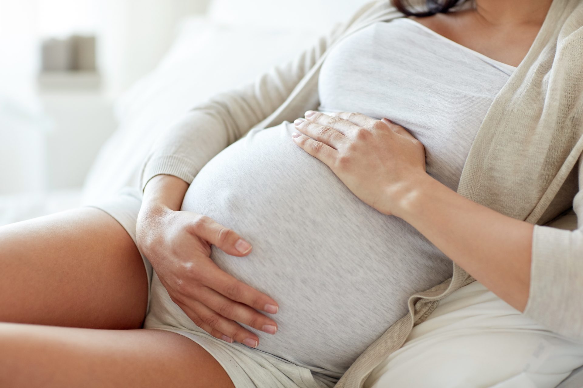 Close-up of a pregnant woman sitting comfortably on a couch with both hands resting on her belly.